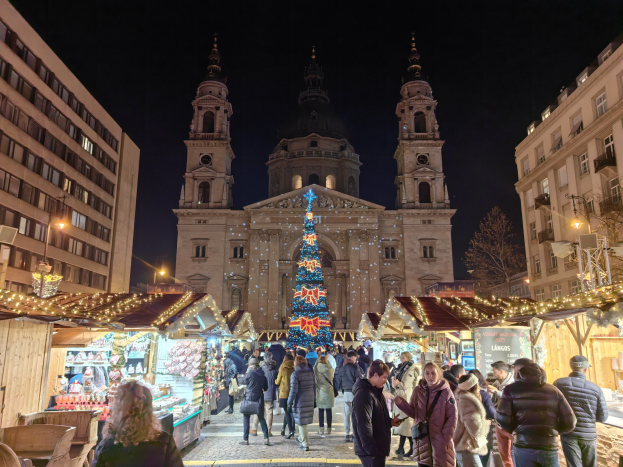 Weihnachtsmarkt vor einer Kirche bei Nacht mit festlich geschmückten Ständen, Menschen, Gebäuden, Laternen, Bäumen und einem sternenklaren Himmel.