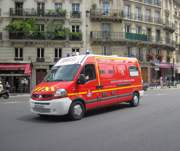 Ein Rettungswagen steht auf der Straße vor einem Gebäude mit Fenstern, Geländern und Pflanzen, mit ein paar Menschen und Fahrrädern im Hintergrund.