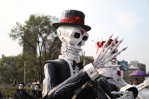 Ein Día de los Muertos-Umzug in Mexiko-Stadt mit Menschen in Kostümen und Schirmen haltend, Skelette, Bäume, Gebäude, Laternenpfähle und eine bewölkte Wolken, mit Text unten.