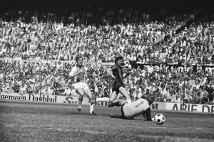 Schwarßerz-Weißerz-Foto von Füßballspielern auf einem Feld mit Zuschauern im Hintergrund und einem Bundesliga 1978-79 Ajax 1-1 Hannover 96-Banner.