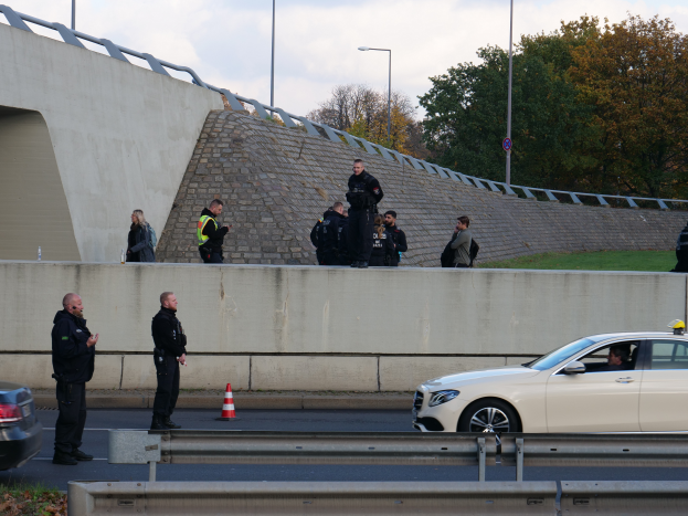 Eine Gruppe von Polizeibeamten steht neben einem Auto auf der Straßenseite, mit Verkehrskegeln, einer Trennwand, Gras, einer Wand, Laternenmästen, Bäumen und einem bewölktem Himmel im Hintergrund.
