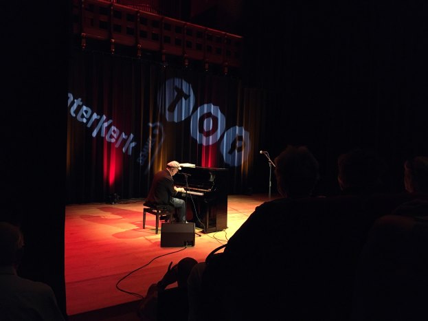 Ein Mann mit Mütze sitzt an einem Piano auf einer schwach beleuchteten Bühne mit einem Mikrofonständer rechts daneben, Kabel auf dem Boden und einem Vorhang mit Text im Hintergrund.
