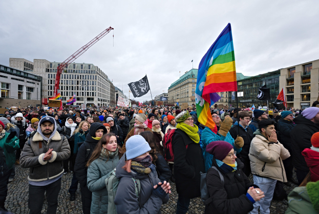 Eine große Gruppe von Menschen steht vor einem Gebäude und hält Fahnen und Schilder mit Text, darunter eines mit der Aufschrift "Lgbtq+ rights march in Berlin", einige tragen Mützen und Taschen, und im Hintergrund sind Gebäude, ein Kran und Wolken zu sehen.