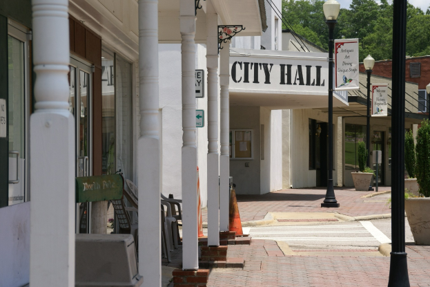 Stadthalle mit umgebender Infrastruktur, einschließlich Straßenlaternen, Schildern, Fahrzeugen, Verkehrskegeln, Stühlen, Zimmerpflanzen, Bäumen und einem bewölkten Himmel.