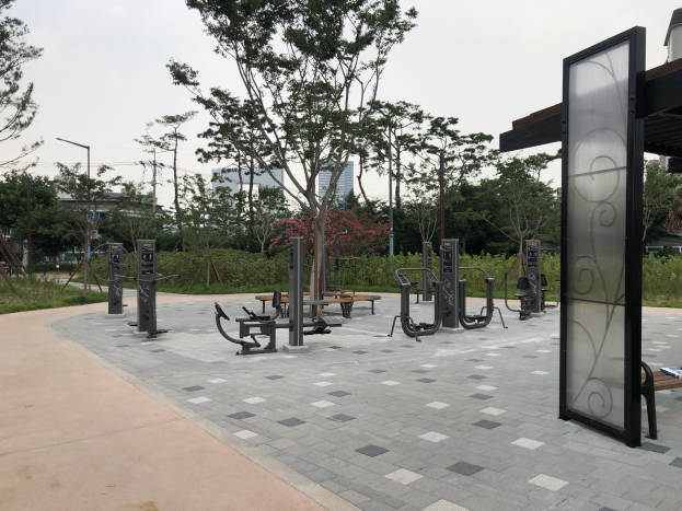 Outdoor-Park mit verschiedenen Fitnessgeräten, Bänken, Bäumen, Pflanzen, Gras, Pfosten, Lichtern, Kabeln und Gebäuden vor einem Himmel-Hintergrund.