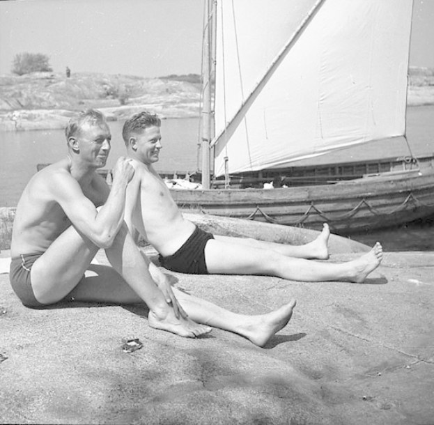 Zwei Männer in Badesachen sitzen auf einem Strand neben einem Segelboot, mit blauem Wasser, grünen Bäumen und einem weißen Himmel im Hintergrund.