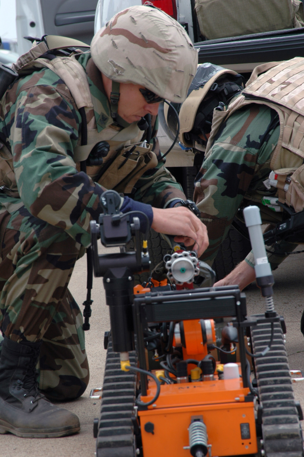 Zwei Männer in Tarnuniformen und Helmen arbeiten an einem kleinen Roboter auf dem Boden, mit Fahrzeugen und Taschen im Hintergrund, was eine militärische Umgebung vermuten lässt.