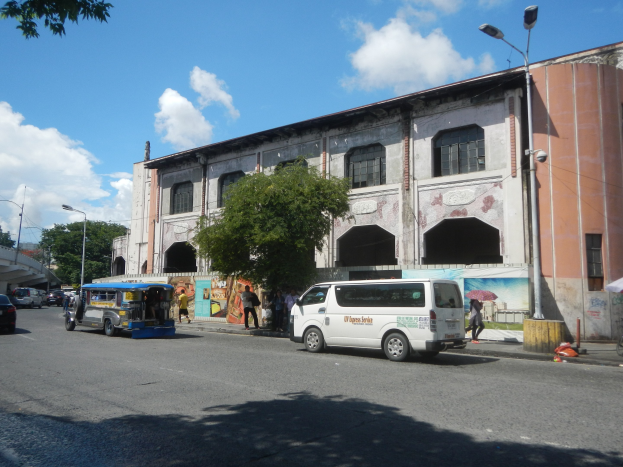 Eine belebte Stadtstraße mit Fahrzeugen, Fußgängern, Bäumen, Laternen, einem großen Gebäude mit einer Wandmalerei, einer fernen Brücke und einem bewölkten Himmel.