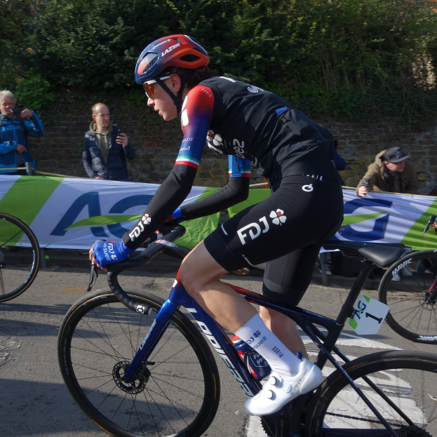 Die professionelle Radrennfahrerin Katie Archibald fährt auf der Straße ein Fahrrad, trägt einen Helm, eine Brille, Handschuhe und Schuhe und fährt an einer Menge Menschen vorbei.