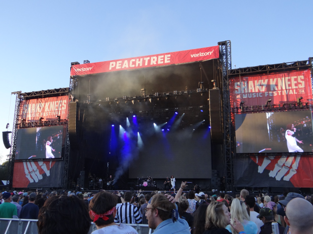 Eine große Menschenmenge steht vor einer hell erleuchteten Bühne auf dem Shaky Knees Music Festival, wo Musiker mit Instrumenten und Bannern hinter ihnen spielen, unter einem klaren blauen Himmel.