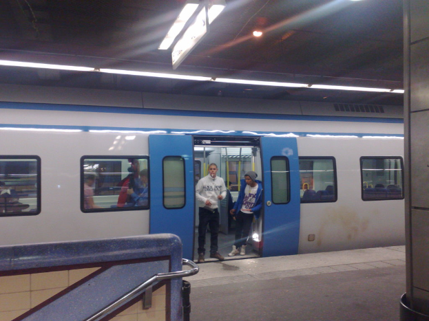 Ein Bahnhofsgebäude mit einem Zug auf den Schienen, zwei Personen an der Zugtür, eine Gruppe von Menschen im Inneren, Lampen oben und ein Geländer links unten.
