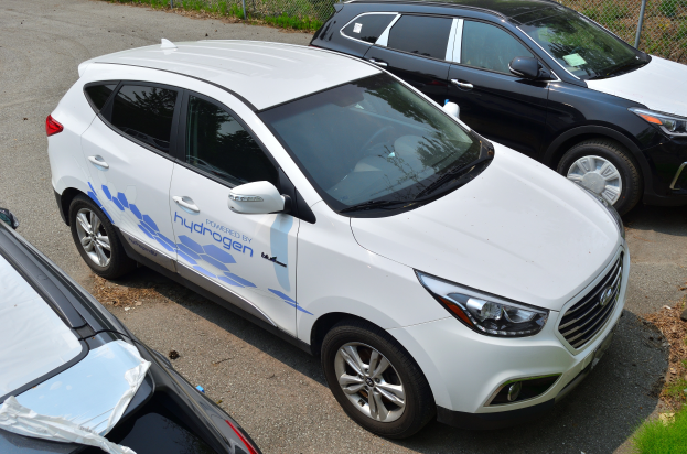 Drei Autos nebeneinander auf einem Parkplatz geparkt, mit einem Maschendrahtzaun und Gras im Hintergrund, eines zeigt den Text Wasserstoff-betriebenes Fahrzeug.