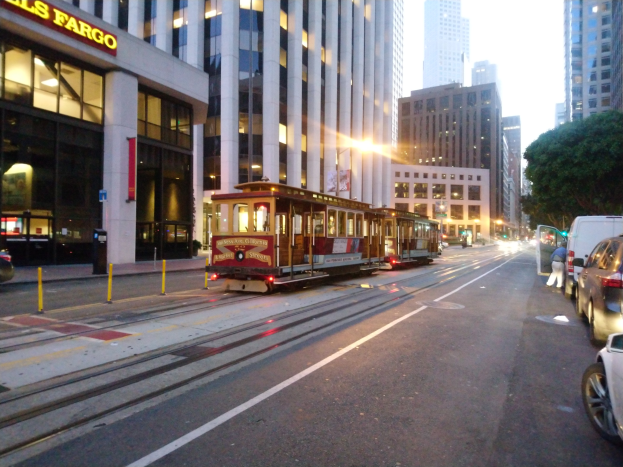 Eine Seilbahn fährt an einer Straße in San Francisco entlang, mit Fahrzeugen, einem Fußgänger und einem geparkten Van im Vordergrund.