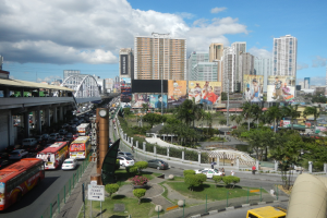 Eine belebte Stadtstraße mit Bussen, Autos und anderen Fahrzeugen, umgeben von Bäumen, Gebäuden, einer Brücke und einem bewölkten Himmel.