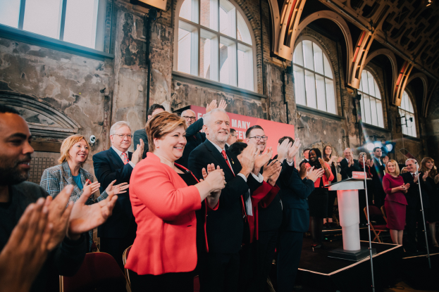 Eine Gruppe von Menschen steht vor einem Publikum und klatscht, mit einem Podium mit Mikrofon auf der rechten Seite und einem Banner im Hintergrund, das "Covid-19 New York City Mayor Bill de Blasio" lautet.