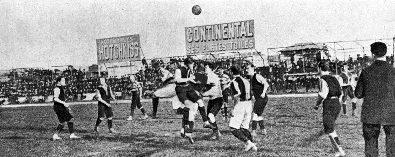 Ein Schwarz-Weiß-Bild einer Gruppe von Männern, die auf einem Feld Fußball spielen, mit einem Ball in der Luft und Zuschauern im Hintergrund, mit Tafeln, die Text über das Spiel anzeigen.