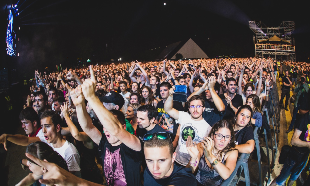 Menschenmenge auf einem Musikfestival mit erhobenen Händen, einige sitzen auf Stühlen und andere stehen, Metallzaun auf der rechten Seite, Gebäude und Lichter im Hintergrund unter einem dunklen Himmel, mit einem Mann und einer Frau im Vordergrund.
