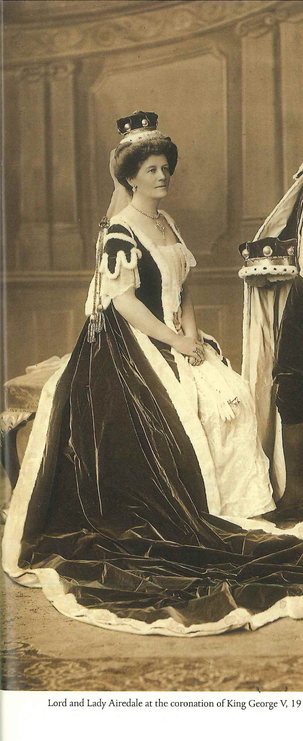 Lady Airedale in einem schwarzen und weißen Kleid vor einer Wand mit einem Objekt rechts daneben und Text unten im Bild.