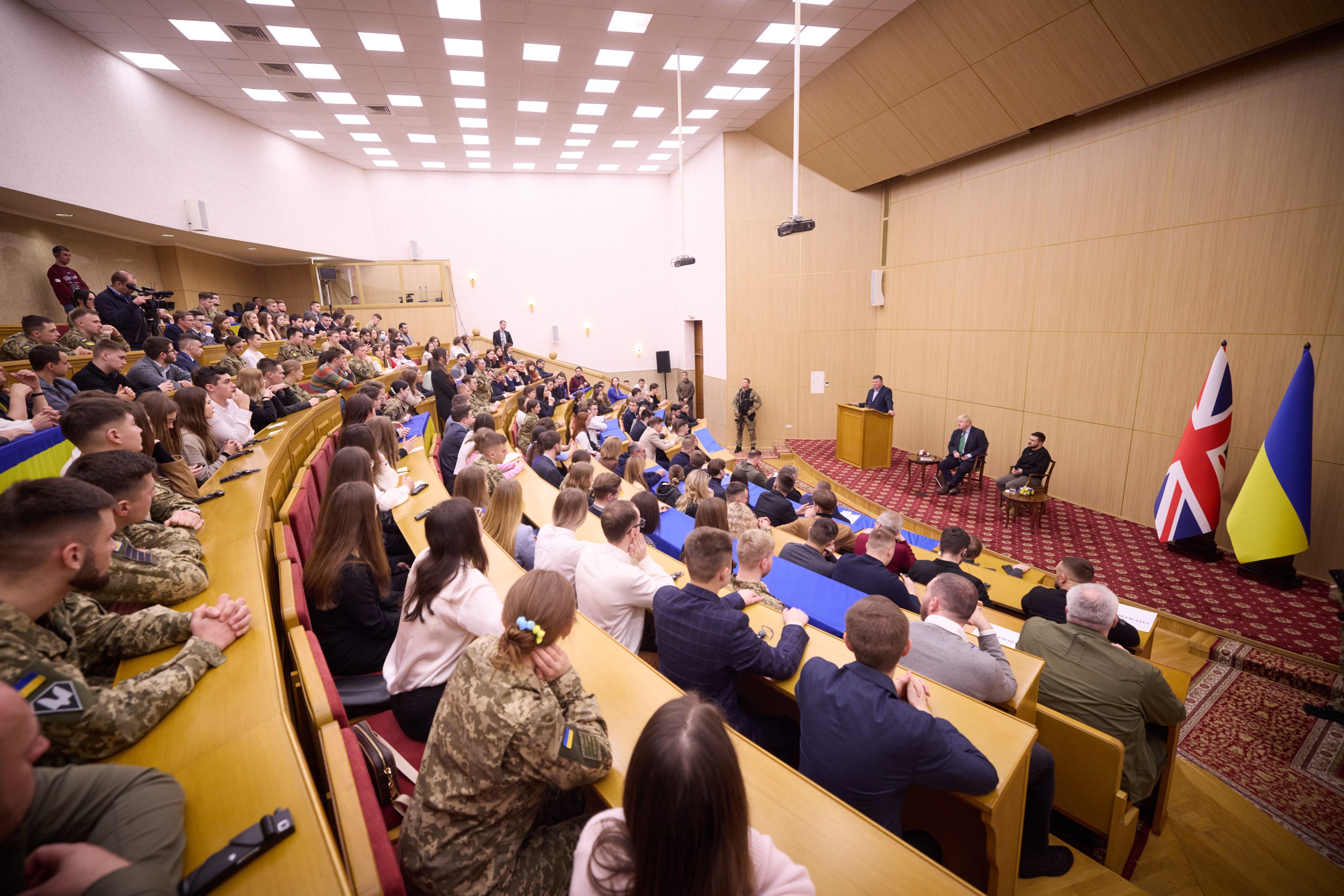 Große Gruppe von Menschen an Tischen in einem Raum mit Flaggen auf der rechten Seite, Papiere und Gegenstände auf den Tischen, einige Stehende vor einem Podium mit Mikrofon und Deckenleuchten und Projektor.