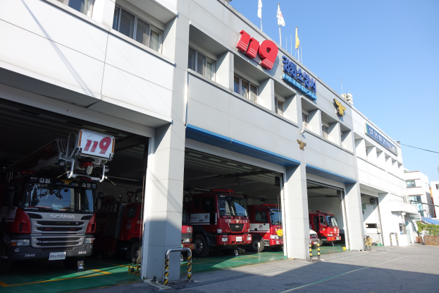 Feuerwache mit mehreren Feuerwehrautos vor der Tür, mit Fenstern, Säulen, Namensschildern und Flaggen im Vordergrund, vor anderen Gebäuden unter einem klaren blauen Himmel.