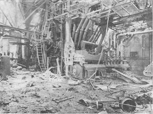 Ein Schwarz-Weiß-Foto eines industriellen Fabrikbodens mit verstreuten Trümmern, darunter Maschinen, Leitern, Pfosten und Drähte, mit sichtbarem Text am unteren Rand.
