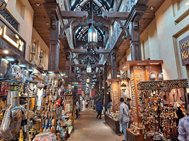 Ein geschäftiges Dubai-Viertel mit Menschen, die zwischen Ständern mit Waren hindurchgehen, Wänden mit Text und einer Decke mit warmem Licht.