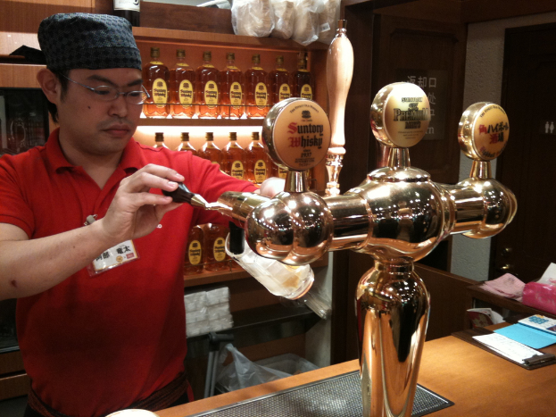 Eine Person mit Mütze und Brille füllt ein Glas mit Bier von einem Automaten auf einem Tisch mit Papieren, mit Regalen voller Alkohol im Hintergrund.