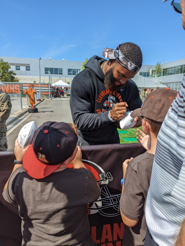 Cleveland Browns Wide Receiver Odell Beckham Jr. signiert Autogramme für Fans vor der Trainingsanlage des Teams, umgeben von Menschen mit Kappen, einem Banner und Gebäuden im Hintergrund.