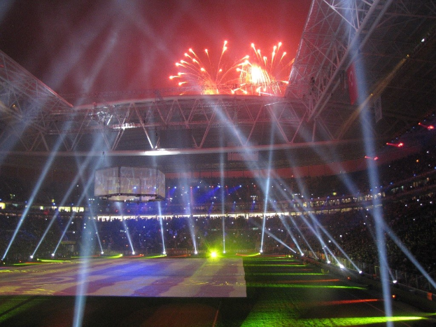 Das Olympische Stadion in London, England, bei Nacht mit hellen Lichtern und Feuerwerken erleuchtet, gefüllt mit Menschen und einem großen Bildschirm im Vordergrund.