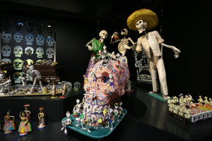 Eine Tag der Toten-Ausstellung im Fine Arts Museum in San Francisco, die Spielzeuge, Skelette und andere Gegenstünde auf Tischen zeigt, mit einer Texttafel im Hintergrund, in einer dunklen und mystischen Atmosph\xare.