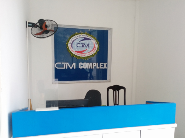 Ein blauer und weißer Empfangstisch mit einem Computer, umgeben von Stühlen und einem Tischventilator, mit einer Tafel an der Wand dahinter, die Text und ein Logo anzeigt, was das Büro von GM Trading Joint in Ho Chi Minh Stadt angibt.