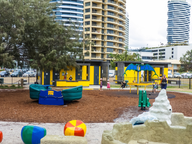 Ein Stadtspielplatz mit Kindern auf der Ausrüstung, umgeben von Bäumen und hohen Gebäuden, mit Fahrzeugen auf der Straße und dem Himmel darüber.