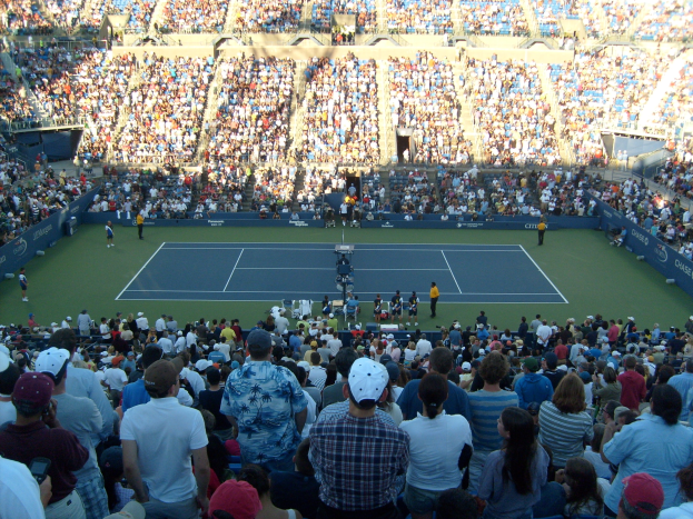 Eine große Menschenmenge schaut einem Tennisspiel in einem vollen Stadion zu, mit Spielern auf dem Platz und Zuschauern in den Rängen.