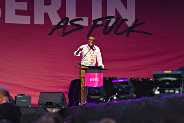 Ein Mann in einem weißen Hemd und braunen Hosen steht auf einer Bühne und hält ein Mikrofon, während er eine Menge anspricht. Im Hintergrund ist ein pinkfarbenes Banner mit der Aufschrift 'Berlin as Fuck' zu sehen.