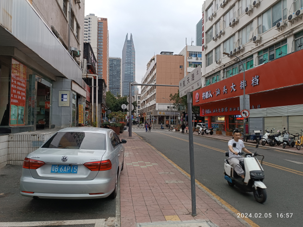 Ein Mann fährt mit einem Roller eine Straße entlang, die von hohen Gebäuden gesäumt ist, mit Menschen auf den Gehwegen und Fahrzeugen auf der Straße, unter einem klaren blauen Himmel mit Bäumen im Hintergrund.