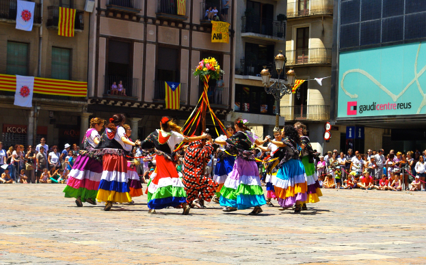 Eine Gruppe von Frauen in bunten Kleidern, die auf der Straße tanzen, umgeben von einer Menge von Menschen, mit Gebäuden, Gelöndern, Balkonen, Flaggen, einer Laterne und einer Tafel mit Schrift im Hintergrund.