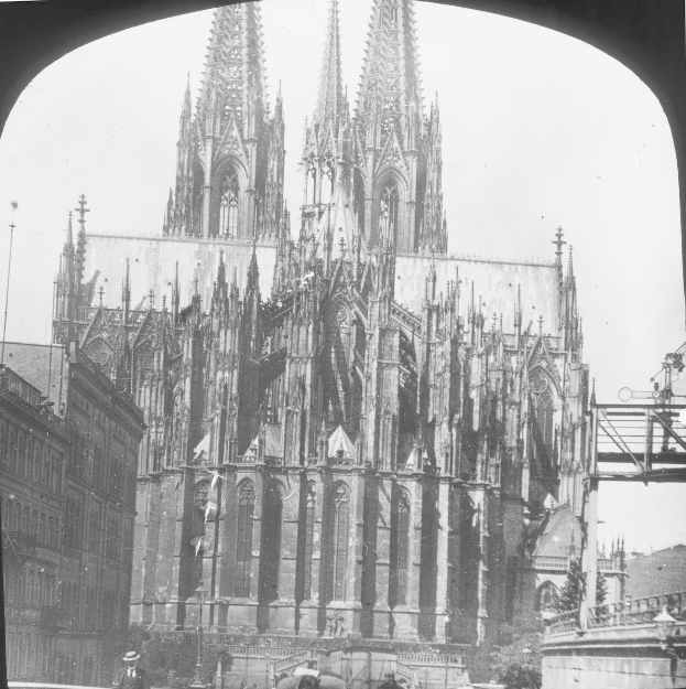 Ein Schwarz-Weiß-Foto des Kölner Doms in Köln, Deutschland, mit einem hohen Turm, umgeben von anderen Gebäuden und Menschen, die auf der Straße davor gehen, mit einer Brücke auf der rechten Seite.