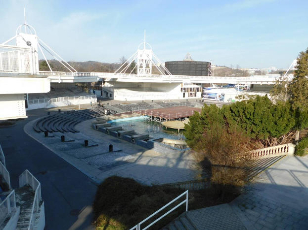 Ein Blick auf das Olympiastadion von einem hohen Standpunkt aus, mit verschiedenen Objekten und Grün im Vordergrund und einem bewölkten Himmel im Hintergrund.