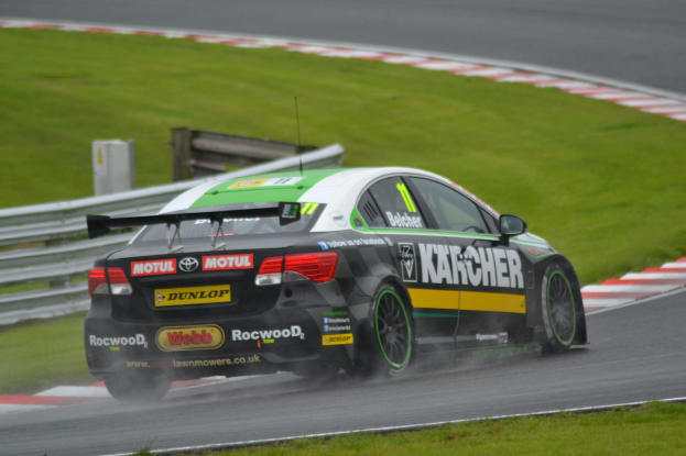 Ein Auto mit Text an der Seite auf einer regennassen Rennstrecke fahrend, umgeben von Gras, mit einem Geländer und einer hölzernen Struktur im Hintergrund.