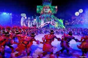 Eine Gruppe von Menschen in bunten Kostümen tanzt auf einer Bühne vor einem sitzenden Publikum, mit Lichtern, Pfählen, Ballons und einem Bildschirm, der eine Person in der Hintergrund zeigt, während des Karnevals in Rio de Janeiro 2014.