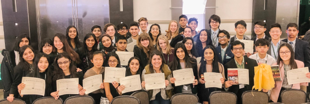 Gruppe von Menschen mit Urkunden bei einer Abschlussfeier in einem Raum mit Stühlen und einer Wand im Hintergrund.