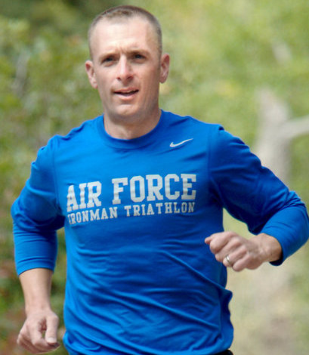 Ein Mann in einem blauen Hemd läuft entschlossen auf einem von Bäumen umgebenen Pfad, der wie ein Luftwaffen-Triathlet aussieht.