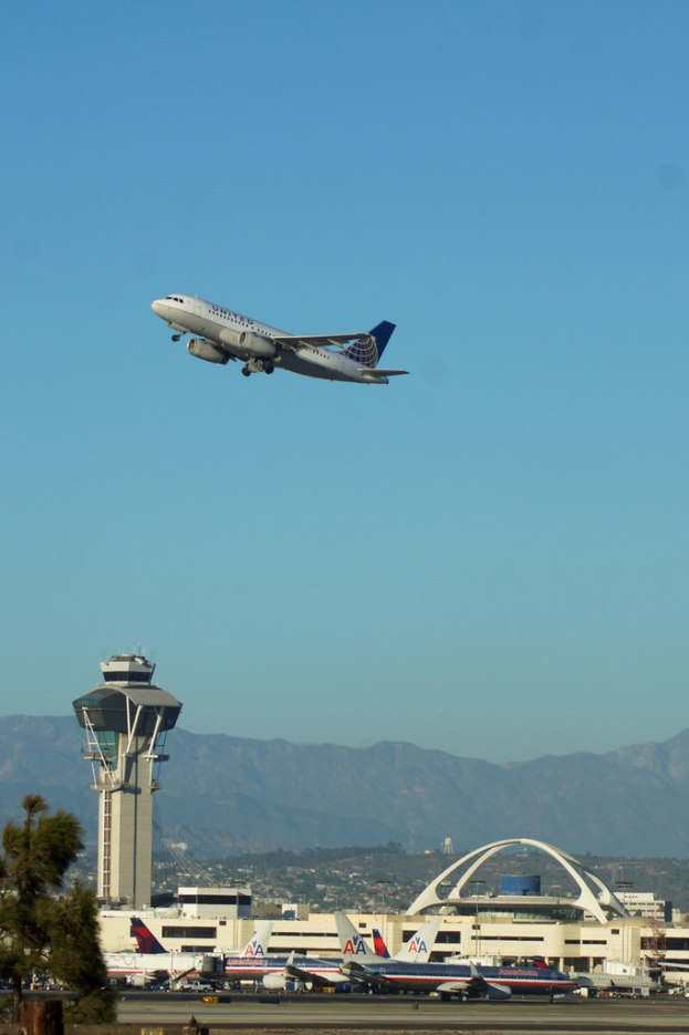 Ein Flughafenblick mit einer weißen Kontrollturm im Vordergrund, geparkten Flugzeugen am Boden, entfernten Gebäuden und einem Flugzeug am Himmel.