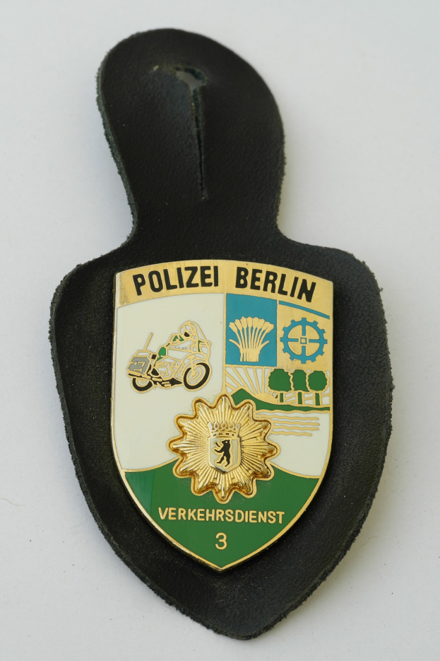 Ein schildförmiges Polizeisiegel mit 'Polizei Berlin' in schwarzer Schrift auf einem blauen Hintergrund, das ein weißes Sternchen und einen weißen Rand hat und auf einer Oberfläche liegt.