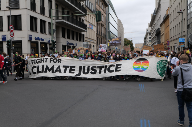 Eine Gruppe von Menschen marschiert mit einem Banner, auf dem "Kampf für Klimagerechtigkeit" steht, eine Straße entlang, mit Gebäuden, Schildern, Ampeln und Bäumen an der Straße unter einem bewölkten Himmel.
