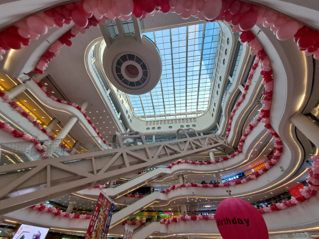 Ein Einkaufszentrum-Innenraum, der für den Valentinstag mit pinken und weißen Ballons, Rolltreppen, Geländern, Texttafeln, Lichtern und einer Glasdecke dekoriert ist.