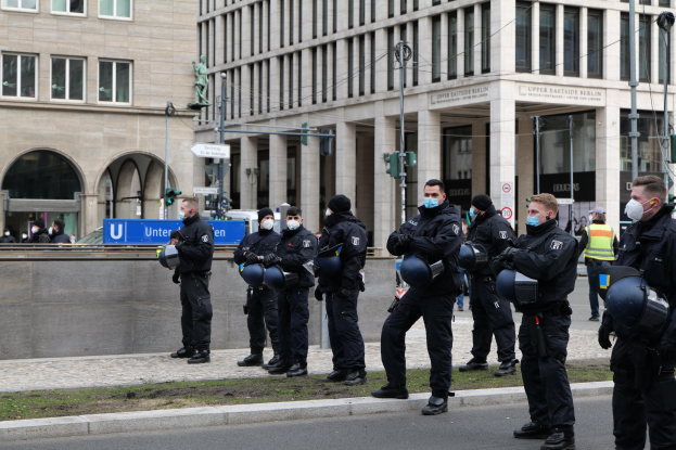 Eine Gruppe von Polizisten in schwarzen Uniformen und Masken steht vor einem Glasfenstergebäude mit Säulen in Berlin, Deutschland, einige halten Helme, mit Laternenmasten, Verkehrsampeln, Texttafeln, einer Statue, anderen Menschen und grasbewachsenem Boden im Hintergrund.
