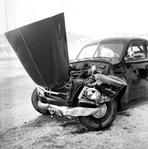 Ein Schwarz-Weiß-Foto eines schwer beschädigten Autos am Straßenrand, mit eingedellter Motorhaube, zersplitterten Fenstern und einer offenen Fahrertür, die den Motor freigibt.