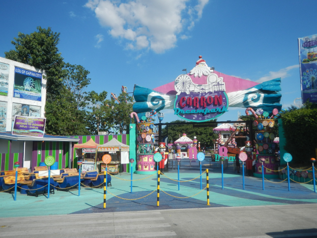 Kirmesattraktion am Parkeingang mit Straße, Umzäunung, Buden, Banner, Bäumen und bewölktem Himmel im Hintergrund.