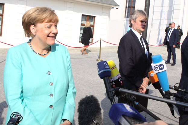 Bundeskanzlerin Angela Merkel beim Sprechen mit Reportern im Freien mit einer Gruppe von Menschen und Equipment vor einem Gebäude mit Fenstern.